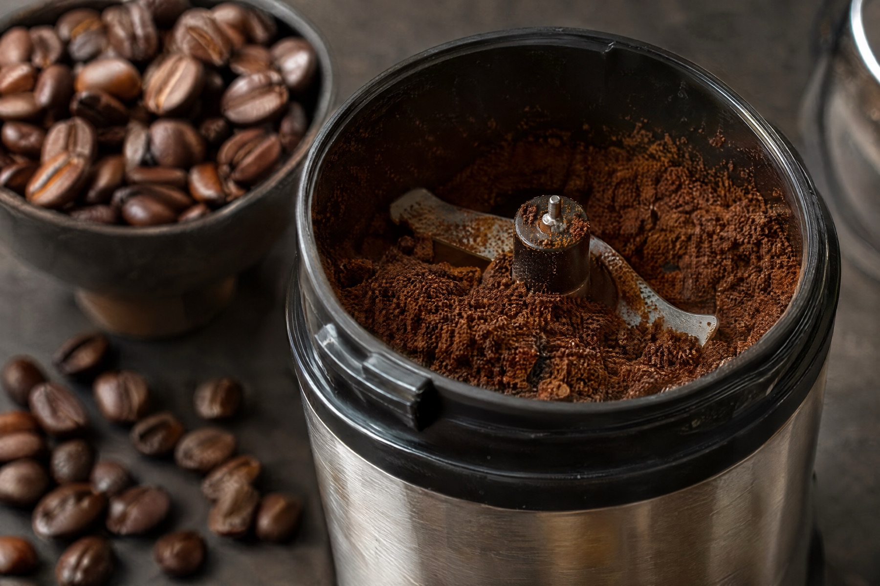 Freshly ground coffee in an open steel grinder, with a bowl of whole roasted beans beside it